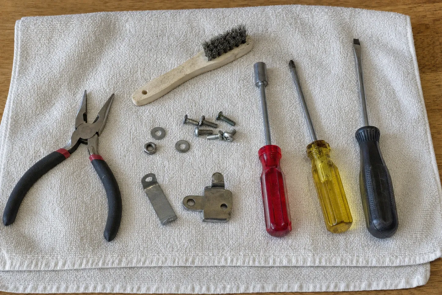 Long-nose pliers, a wire brush, three drivers, two small brackets, washers and screws laid out on a cloth