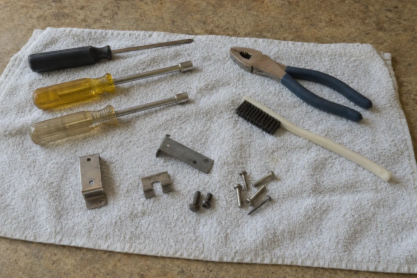 Black Phillips driver and two yellow-handled nut drivers beside an angle bracket, a drilled steel strip, a small U-clip, machine and self-tapping screws, slip-joint pliers and a white-handled wire brush on a towel